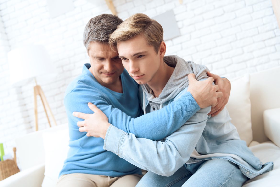 Father hugs son while sitting with him at home on the couch. Father and son forged family relationships together.