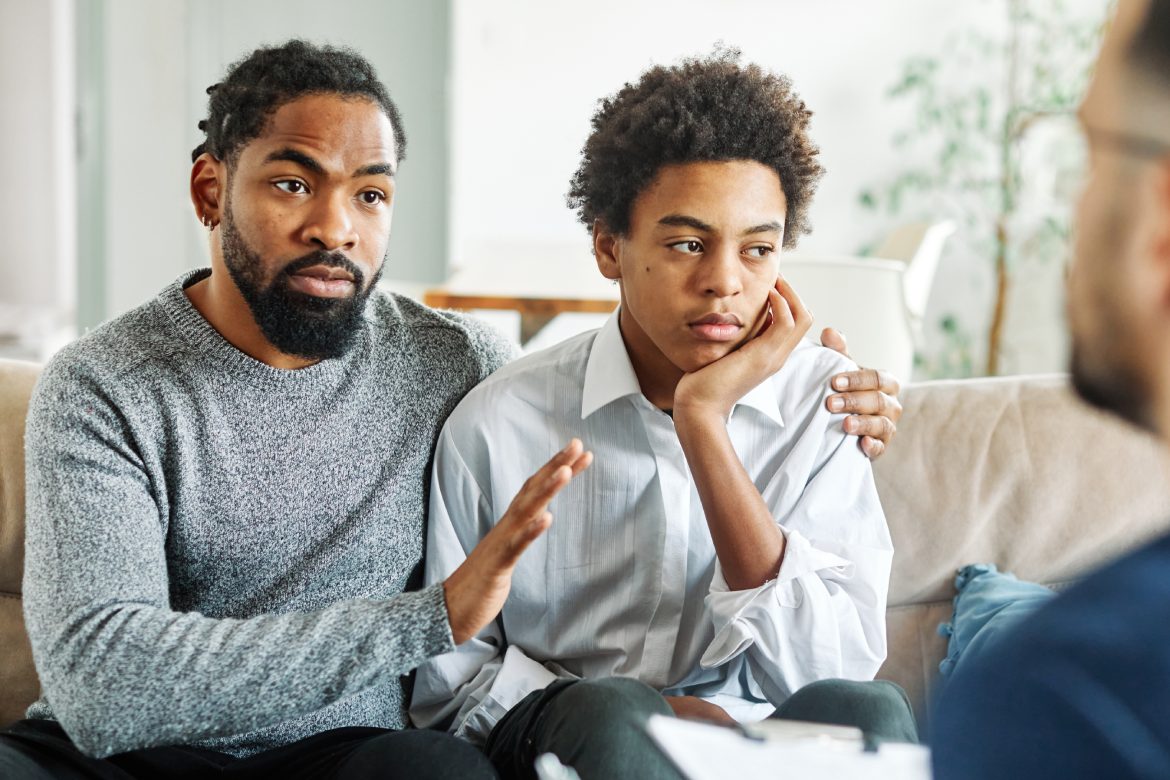 Father with his teenage son at meeting with social worker, psychologist discussing mental health family sitting on sofa in psychotherapist office