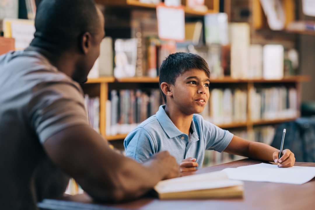 Young school boy having a lesson with his teacher in a library. Elementary school student talking to his tutor and writing in a book. Academic support in a primary school.