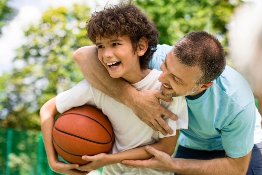 Happy Loving Family. Portrait of laughing dad hugging his curly son from the back, boy holding basket ball